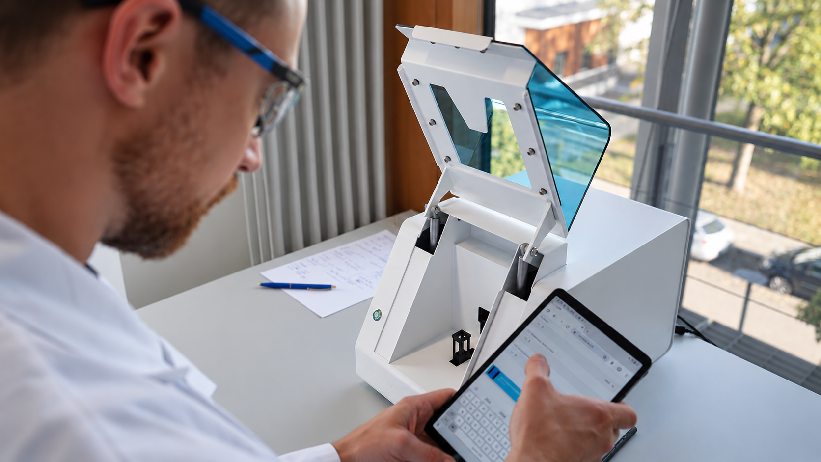Researcher operating Xell from a tablet, with the printer's lid open on a lab bench by the window